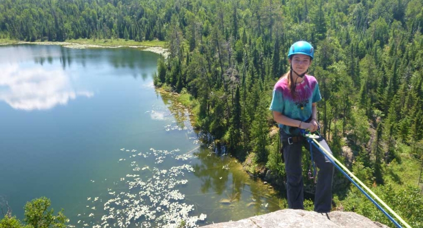 A person wearing safety gear is secured by ropes as they look at the camera and smile while rock climbing. They are high above a body of water and a green forrest. 